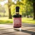 Eine freigestellte Produktaufnahme einer 20cl-Glasflasche mit „TOKO Rote Beeren Likör“ Aufschrift, die auf einem Holztisch im Freien steht. Die Flasche hat einen schwarzen Verschluss und ist mit einer intensiven, tiefroten Flüssigkeit gefüllt. Das weinrote Etikett zeigt eine feine Illustration verschiedener Waldbeeren und trägt die Aufschrift „100% aus Frucht“ sowie „20% Vol.“.